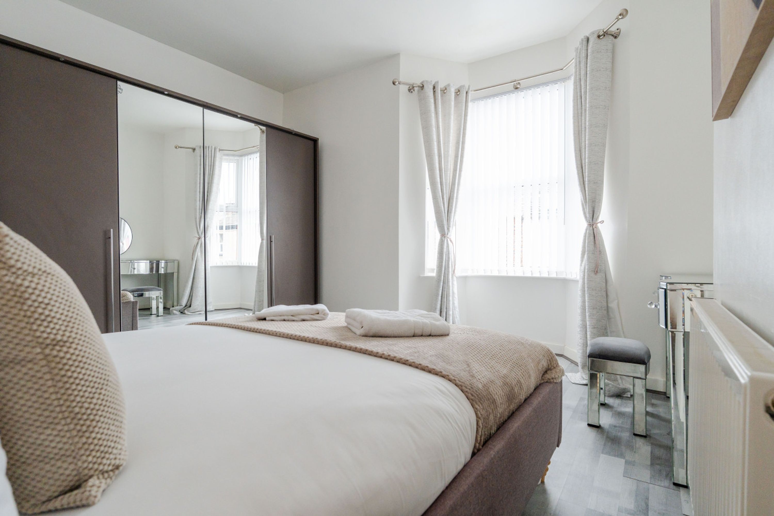 Une chambre à coucher comprenant un lit, une armoire, un miroir, une fenêtre avec des rideaux et un radiateur.