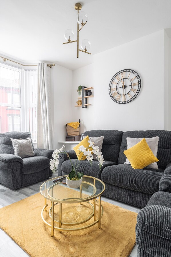 A modern living room with a grey sofa, yellow cushions, a glass coffee table, and a wall clock. - Newly Renovated 4BR House With Lots of Amenities (Bootle)