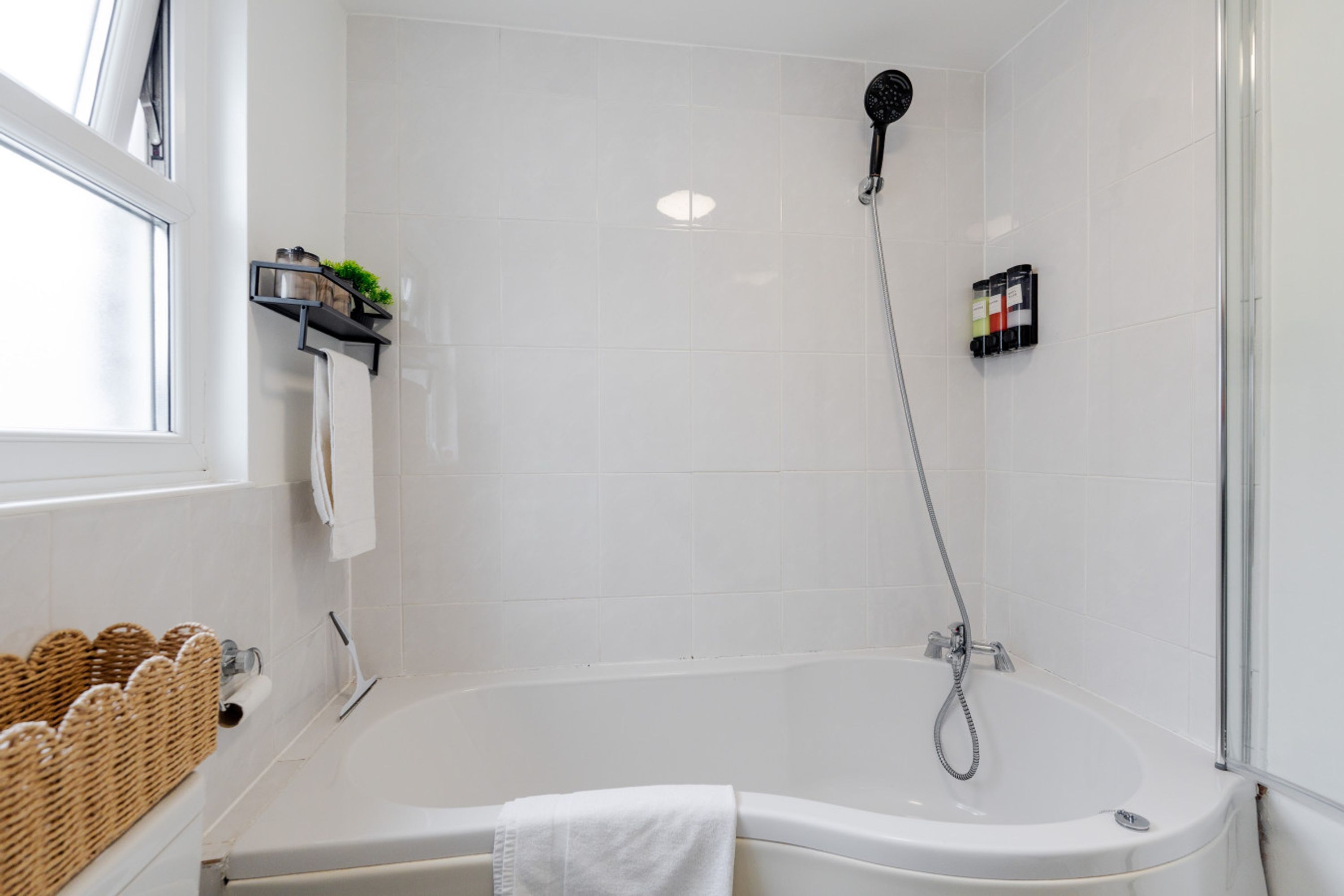 Ein modernes Badezimmer mit einer Badewanne, einer Dusche, weißen Fliesen, einem Fenster und einem Ablagefach mit Toilettenartikeln.
