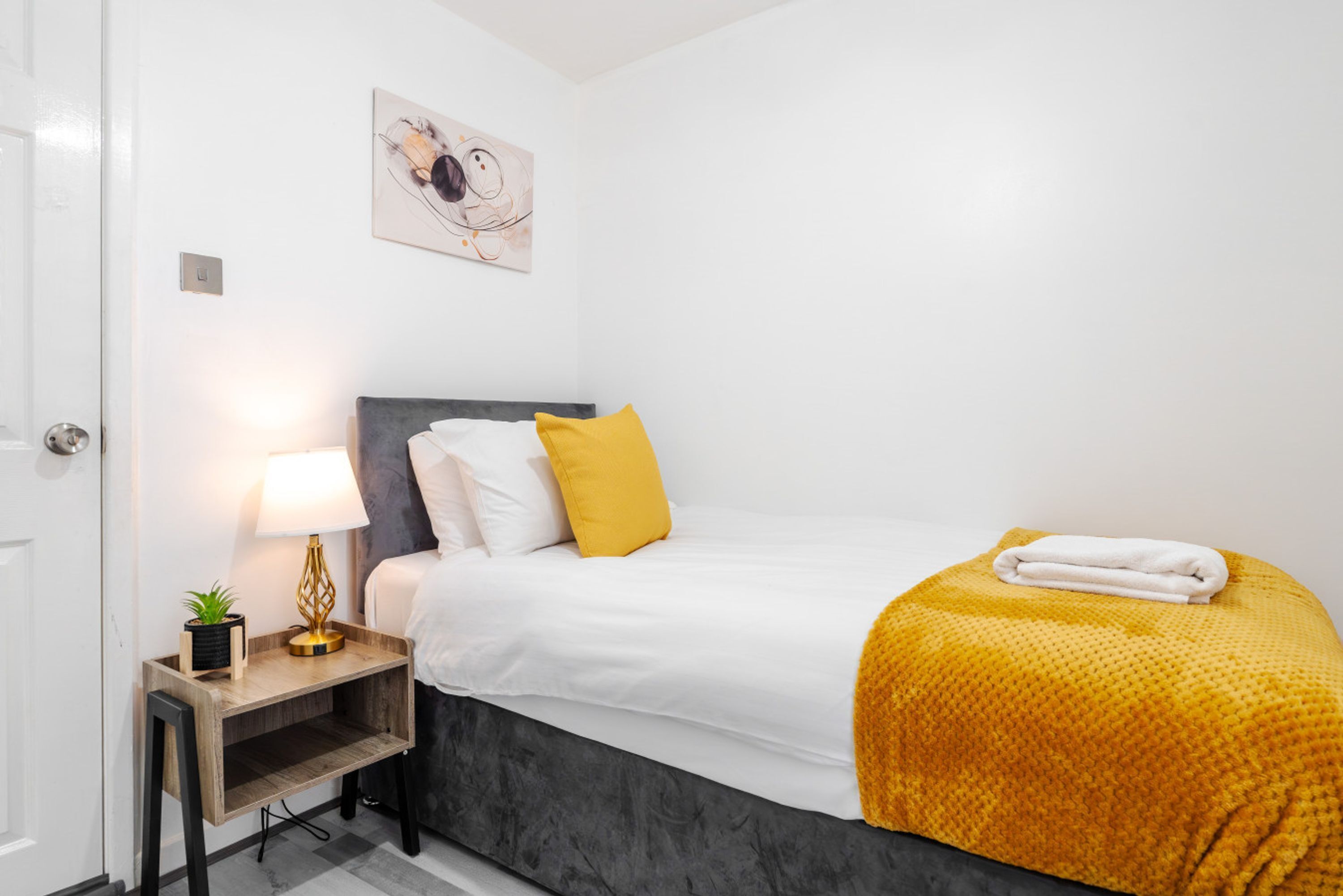 A bedroom with a bed, bedside table, lamp, and artwork on the wall.