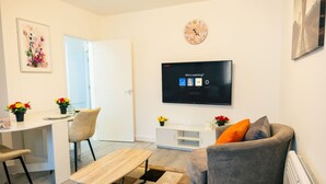 Ein modernes Wohnzimmer mit Flachbildfernseher, Essbereich mit Tisch und Stühlen sowie ein graues Sofa mit orangem Kissen.