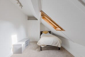 Una habitació petita i blanca amb un sostre inclinat, un llit amb matalàs blanc i almoina groga, un prestatge blanc i un armari petit i blanc.