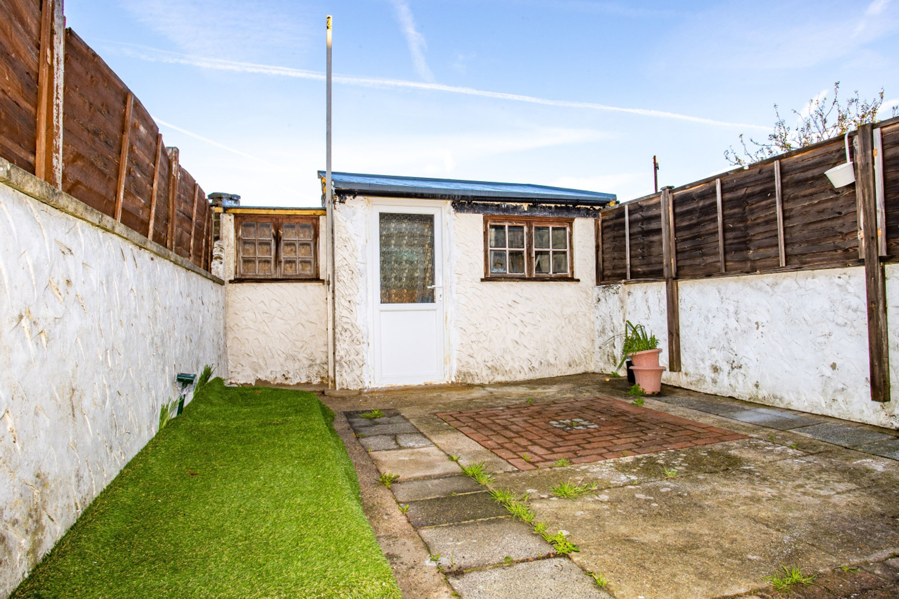 A small, fenced backyard with a white building, a paved area, and a grassy patch.