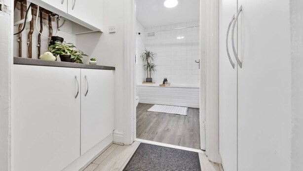 Un pasillo estrecho con armarios blancos, suelo de madera y zona de ducha con pared de azulejos.