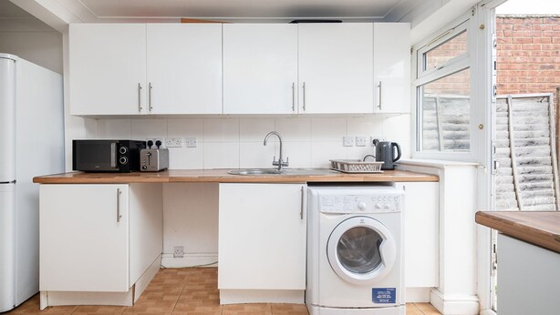 A compact kitchen with white cabinets, a washing machine, a microwave, and a sink. - Family-friendly 3-bed Haven in Northolt w Parking (Northolt)