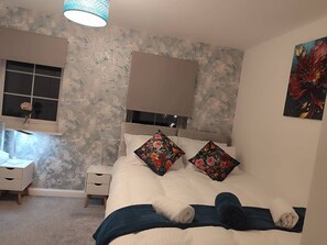 A bedroom with a bed, a nightstand, a window, and a wall with a floral pattern. - Spacious Home for Groups Sleeps 8 (Stoke-on-Trent)
