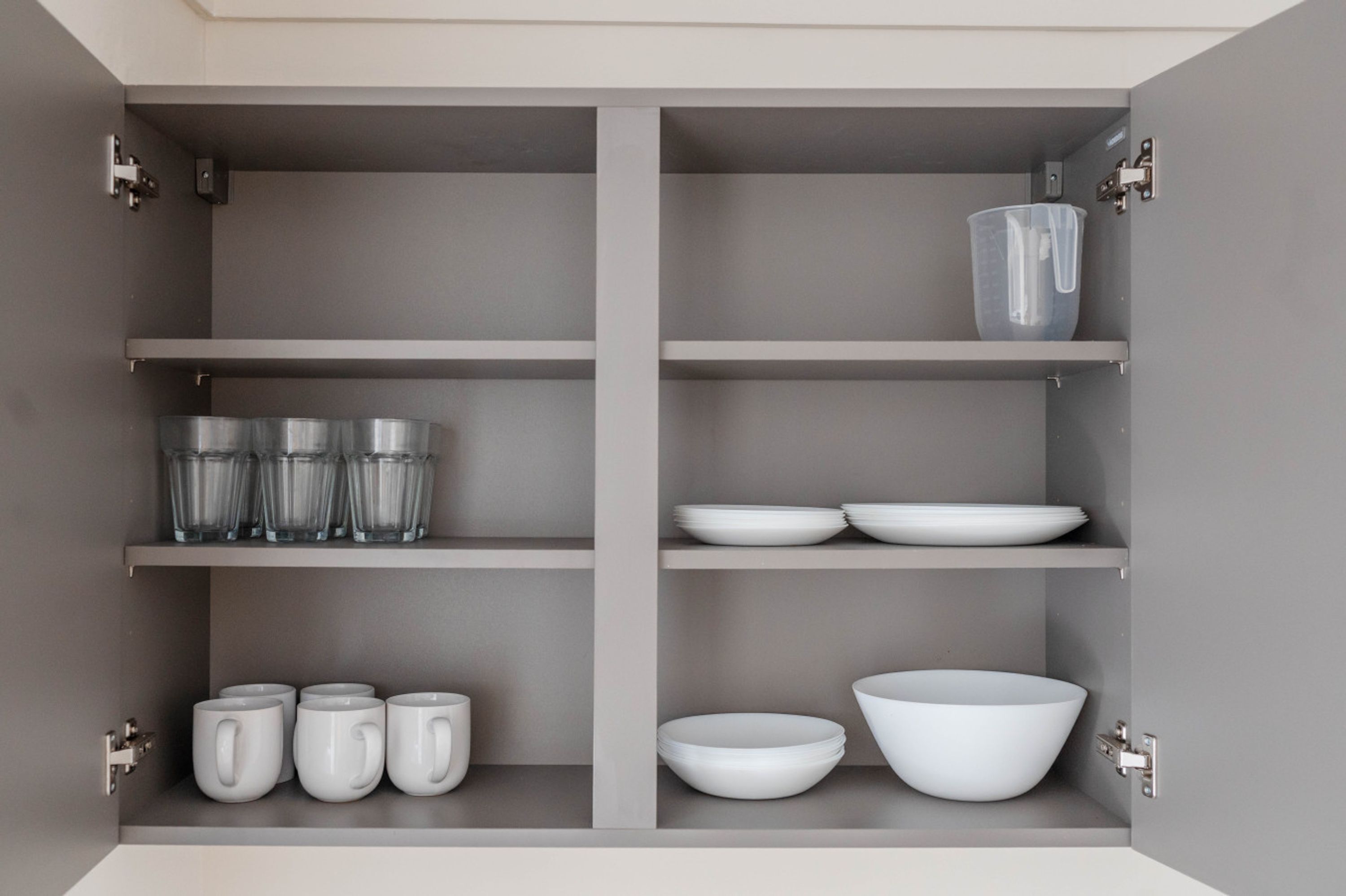 A kitchen cabinet with shelves holding clear glasses, white mugs, white plates, and a large white bowl.