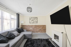 Een woonkamer met een grijze bank, een zwart tapijt, een open haard, een televisie en een metselwerk muur.