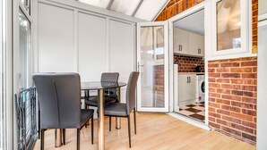 Zona pranzo con tavolo e sedie, cucina visibile attraverso una porta aperta e parete in mattoni.