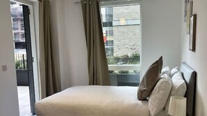 Ein Schlafzimmer mit einem Bett, einem Nachttisch, einer Lampe, einem Fenster mit Vorhängen und Blick nach draußen.