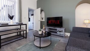 A living room with a television displaying 'NETFLIX,' a sofa, a coffee table with a bottle and glasses, and a dining area with benches. - Stylish 3BR Home With Parking by University (Sunderland)
