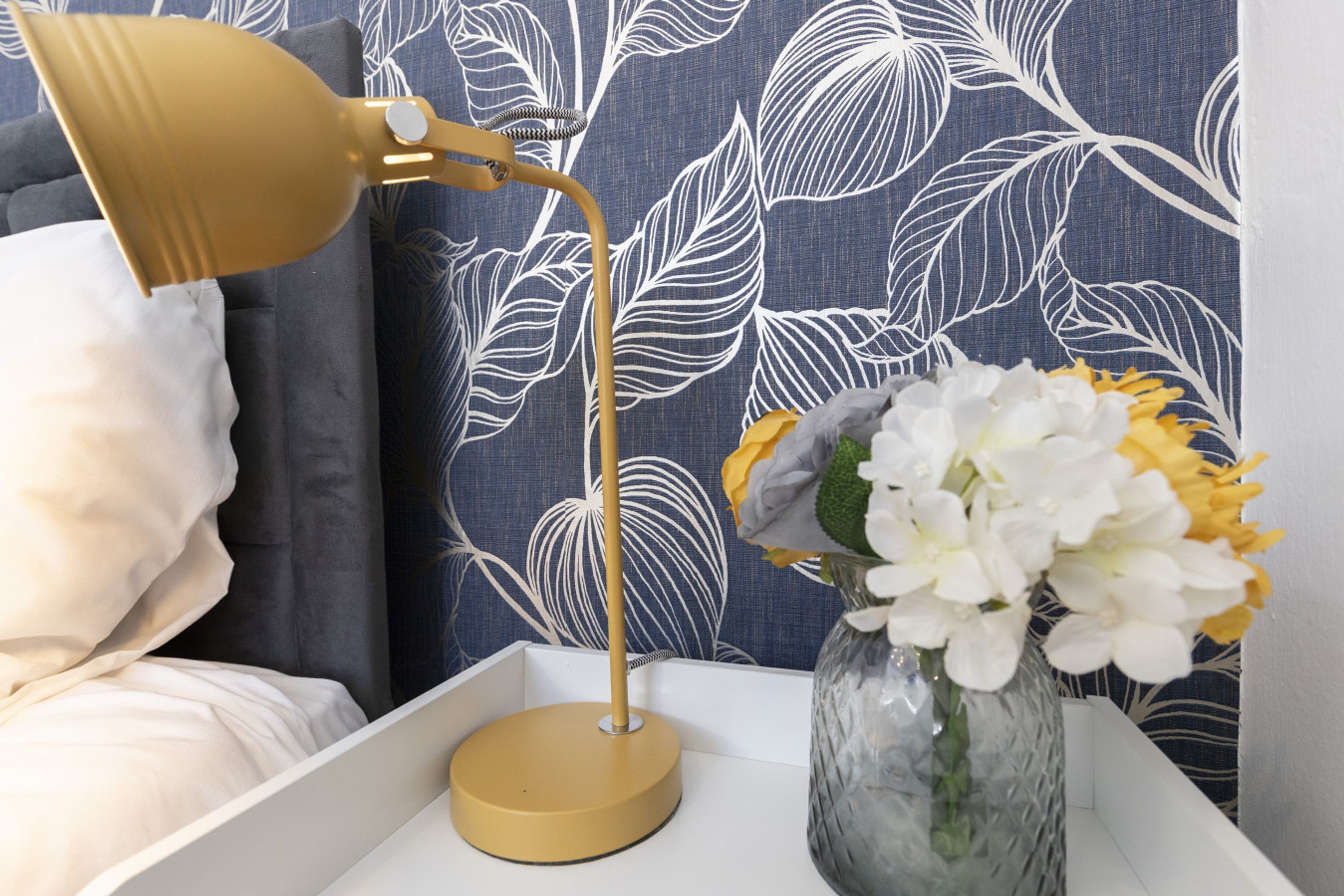 A bedside table with a gold lamp, a vase with flowers, and a blue patterned wall.