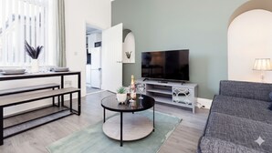 A living room with a sofa, a TV, a dining table, and a kitchen in the background. - Stylish 3BR Home With Parking by University (Sunderland)