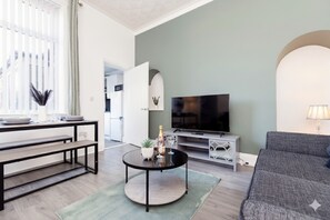 Ein Wohnzimmer mit Sofa, Fernseher, einem Esstisch und einer Küche im Hintergrund.