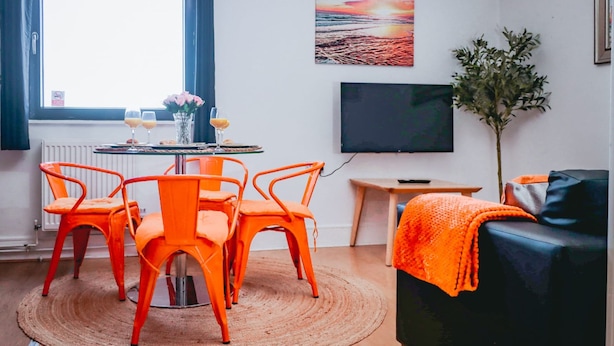 A living room with a round table, orange chairs, a black sofa, a TV, and a plant. - Elizabeth Line Retreat Modern 2BR Flat in Hayes (Hayes)