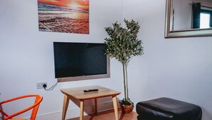 A room with a flat-screen TV mounted on the wall, a small wooden table, a potted plant, a black leather ottoman, and a colorful sunset painting. - Elizabeth Line Retreat Modern 2BR Flat in Hayes (Hayes)