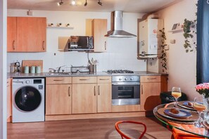A compact kitchen with a washing machine, microwave, stove, oven, and wooden cabinets. - Elizabeth Line Retreat Modern 2BR Flat in Hayes (Hayes)