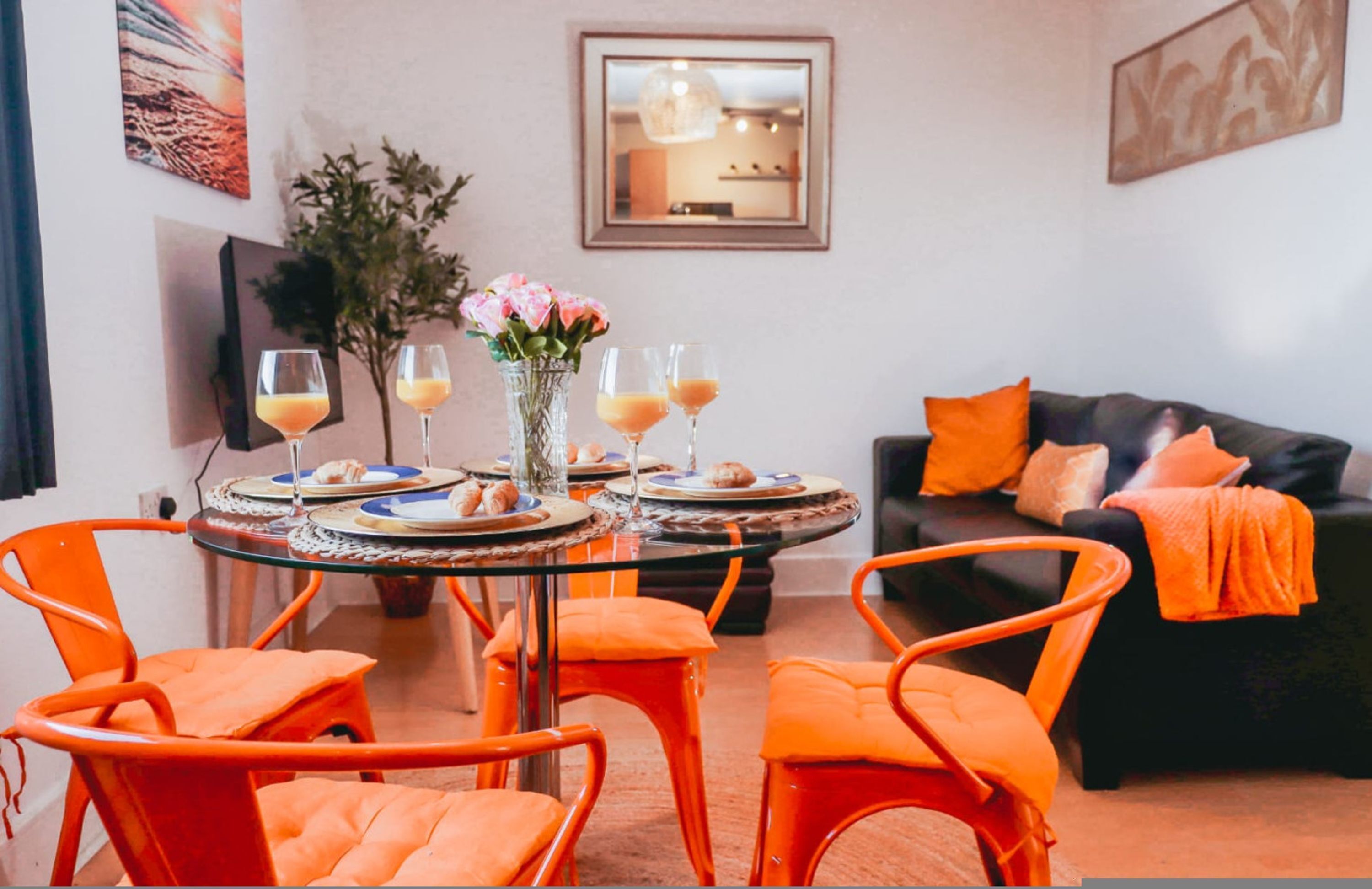Un comedor con una mesa redonda, sillas naranjas y un sofá negro con cojines anaranjados.