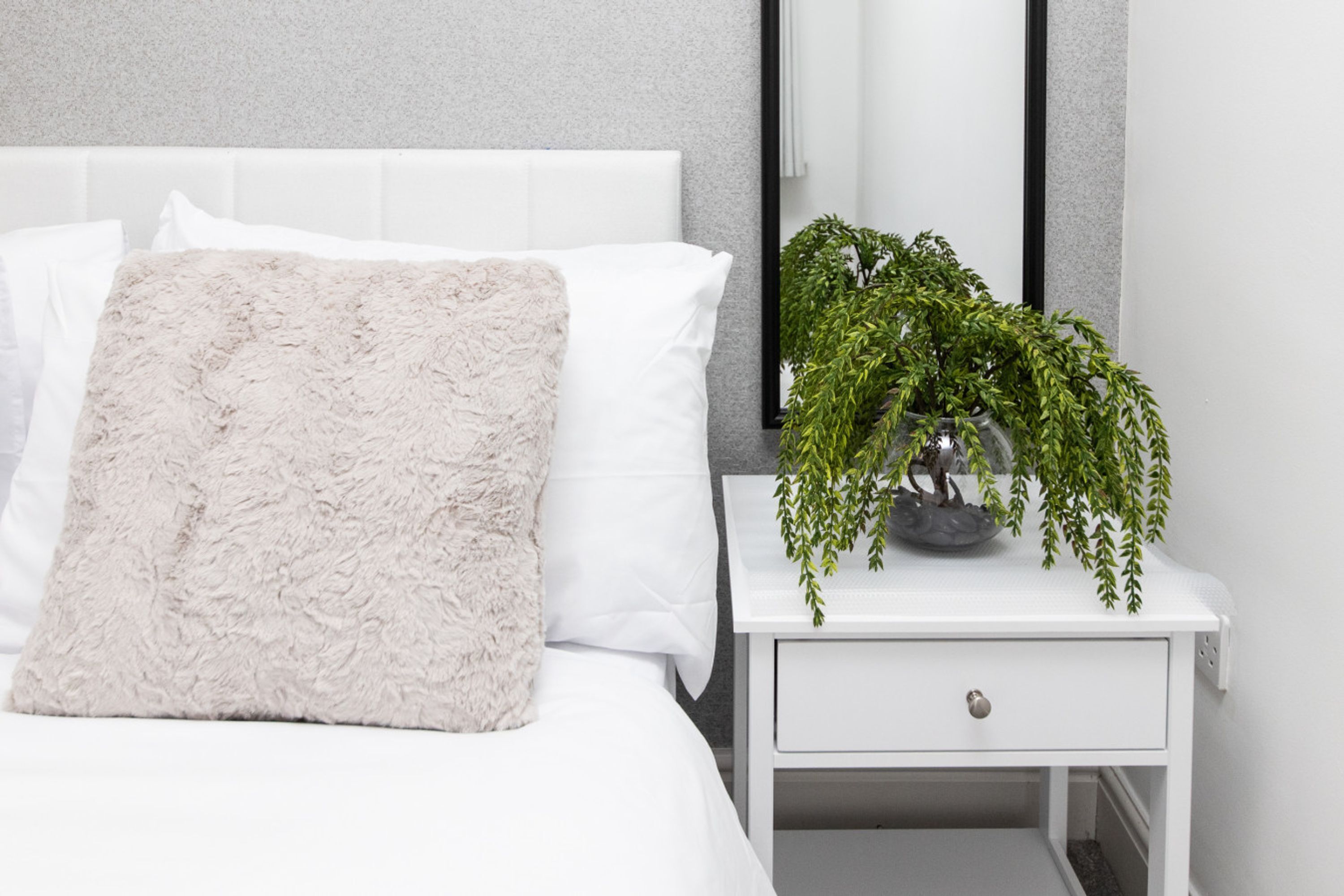 A neatly made bed with a fluffy pillow, a white bedside table with a plant, and a white drawer.