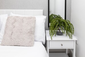 Un lit bien fait, avec un oreiller moelleux, une table de chevet blanche ornée d’une plante et un tiroir blanc.