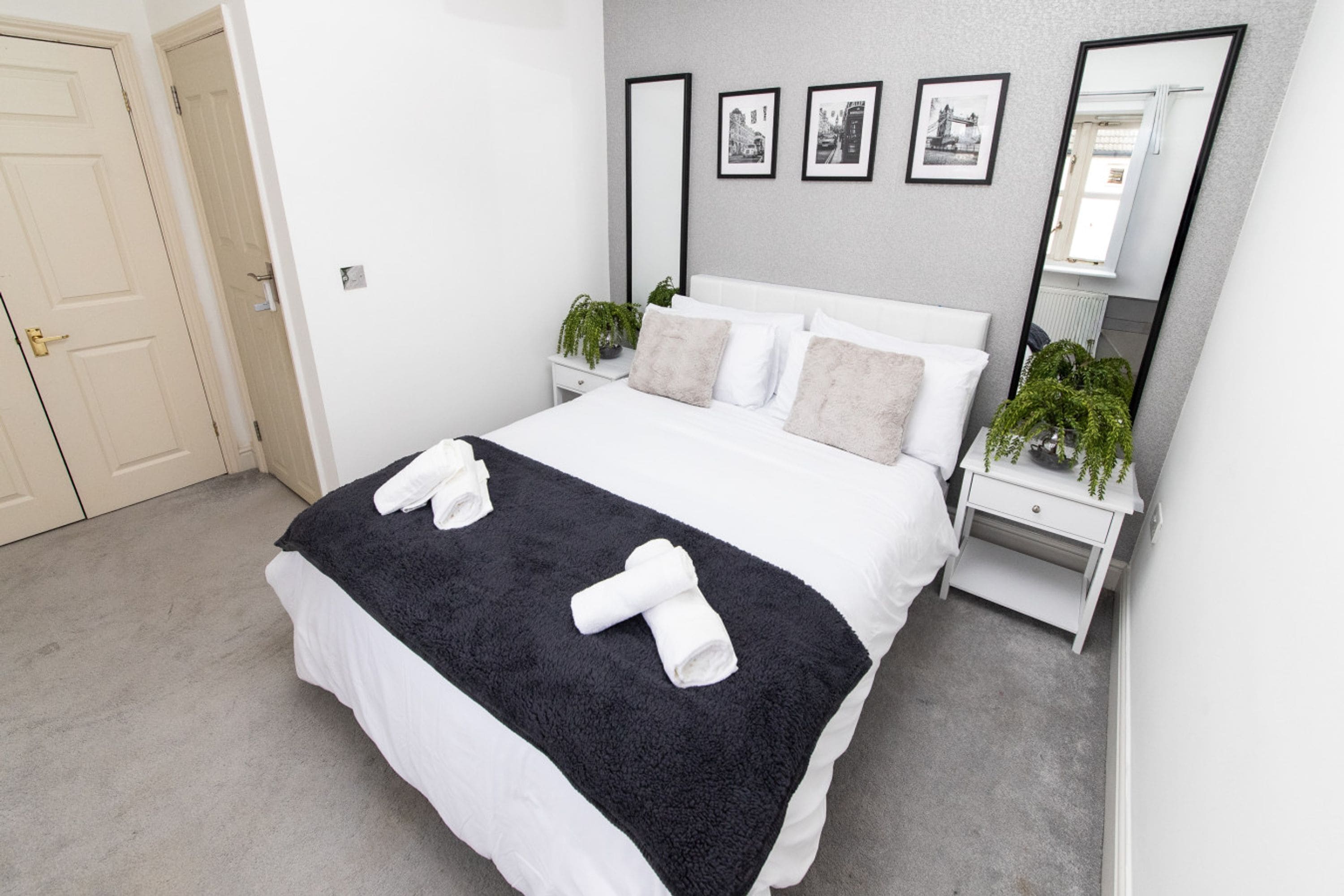 A neatly made bed with a black and white blanket, two bedside tables with plants, a large mirror, and framed pictures on the wall.