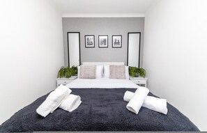 Une chambre à coucher moderne avec un grand lit, deux tables de chevet, un mur d’accent de couleur grise et trois tableaux encadrés.