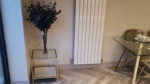 A room with a herringbone floor, a radiator, a glass table with a chair, and a vase with dark foliage. - London 2BR Retreat With Free Parking City Access (London)