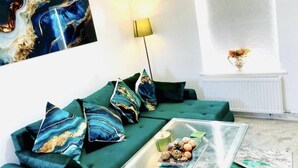 Een woonkamer met een groene bank, decoratieve kussens, een glazen salontafel met lekkernijen, een vloerlamp en een schilderij aan de muur.