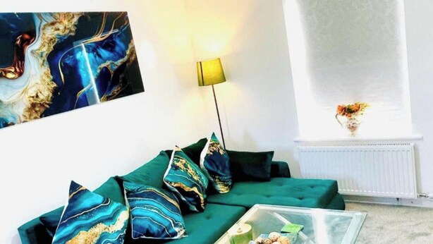 Een woonkamer met een groene bank, decoratieve kussens, een vloerlamp, een vaas met bloemen en een schilderij aan de muur.