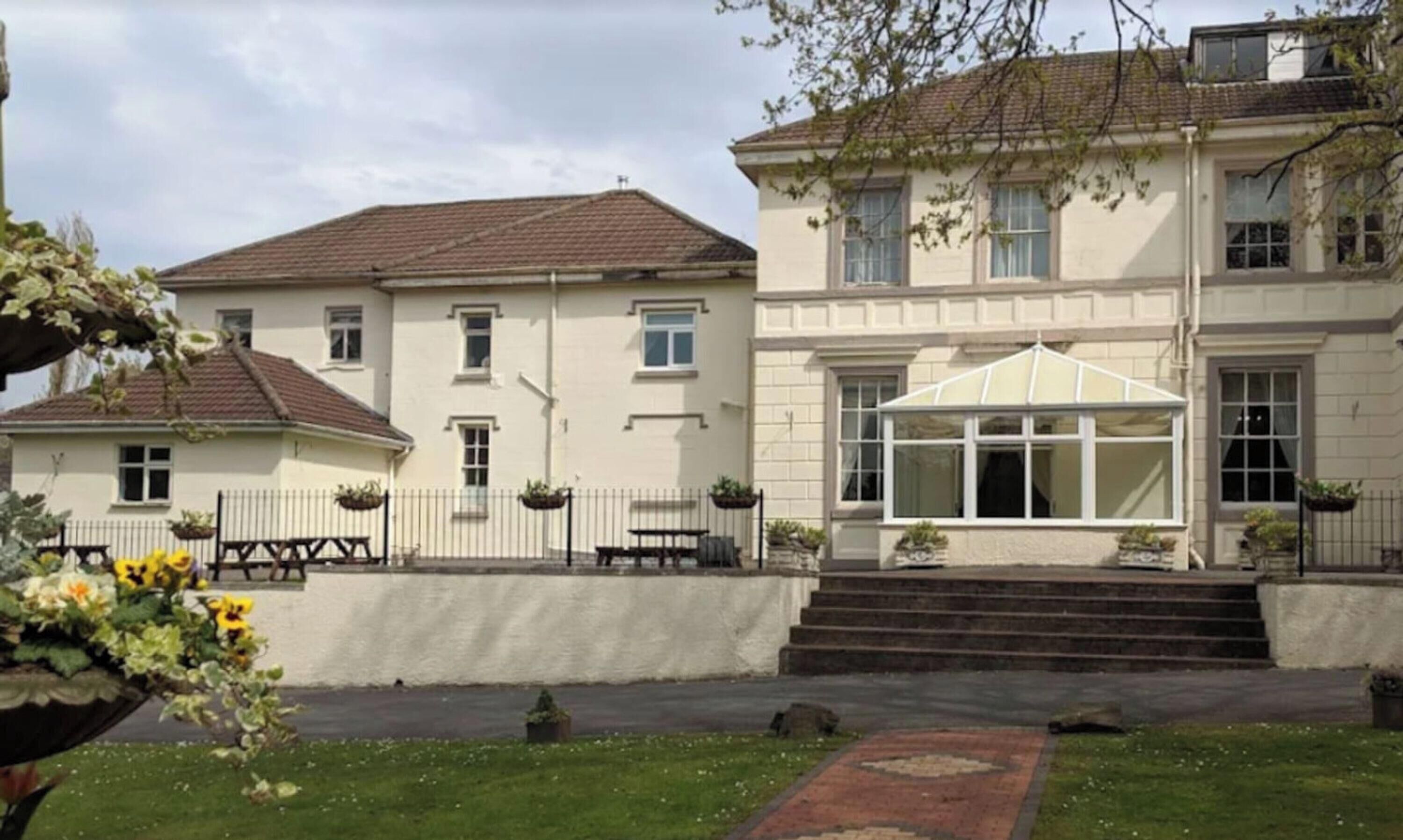 Un edificio a due piani con veranda, vialetto lastricato e piccolo giardino fiorito.