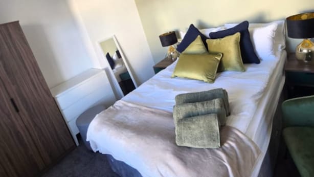 A hotel room with a bed, bedside tables, a mirror, a wardrobe, and a hanging light fixture. - West Midlands Home - 3BR w Free Parking and Wifi (Dudley)