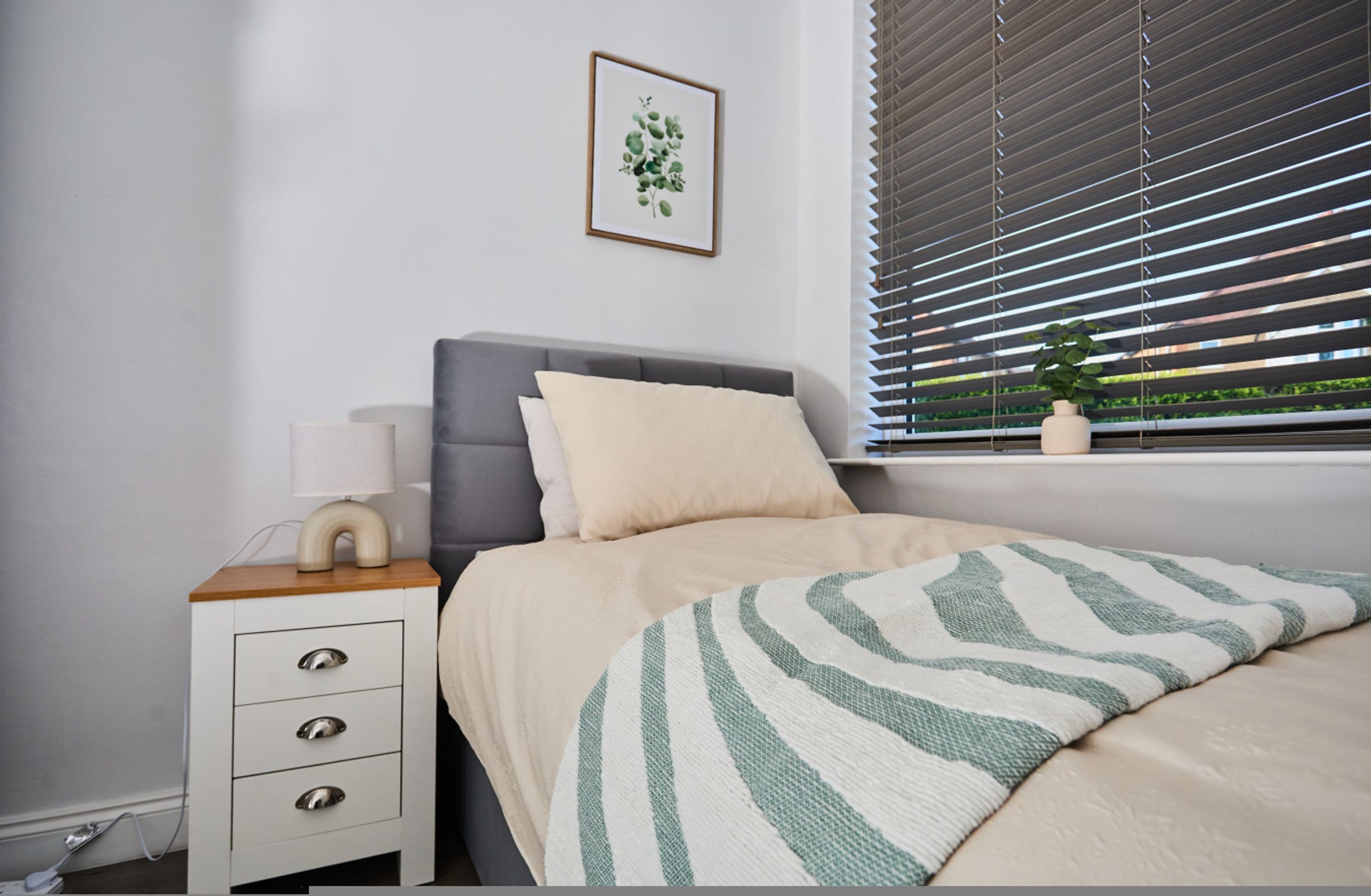 A bedroom with a bed, bedside table, lamp, and a window with blinds.