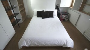 A neatly made bed with white bedding and black pillows, a radiator, a shelf with items, and a window with blinds. - Stylish East London Retreat With Private Terrace Fast Wifi (London)