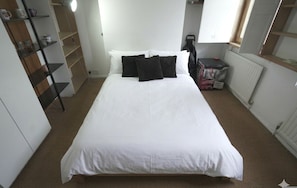 Ein ordentlich bezogenes Bett mit weißer Bettwäsche und schwarzen Kissen, ein Heizkörper, ein Regal mit Gegenständen und ein Fenster mit Jalousien.