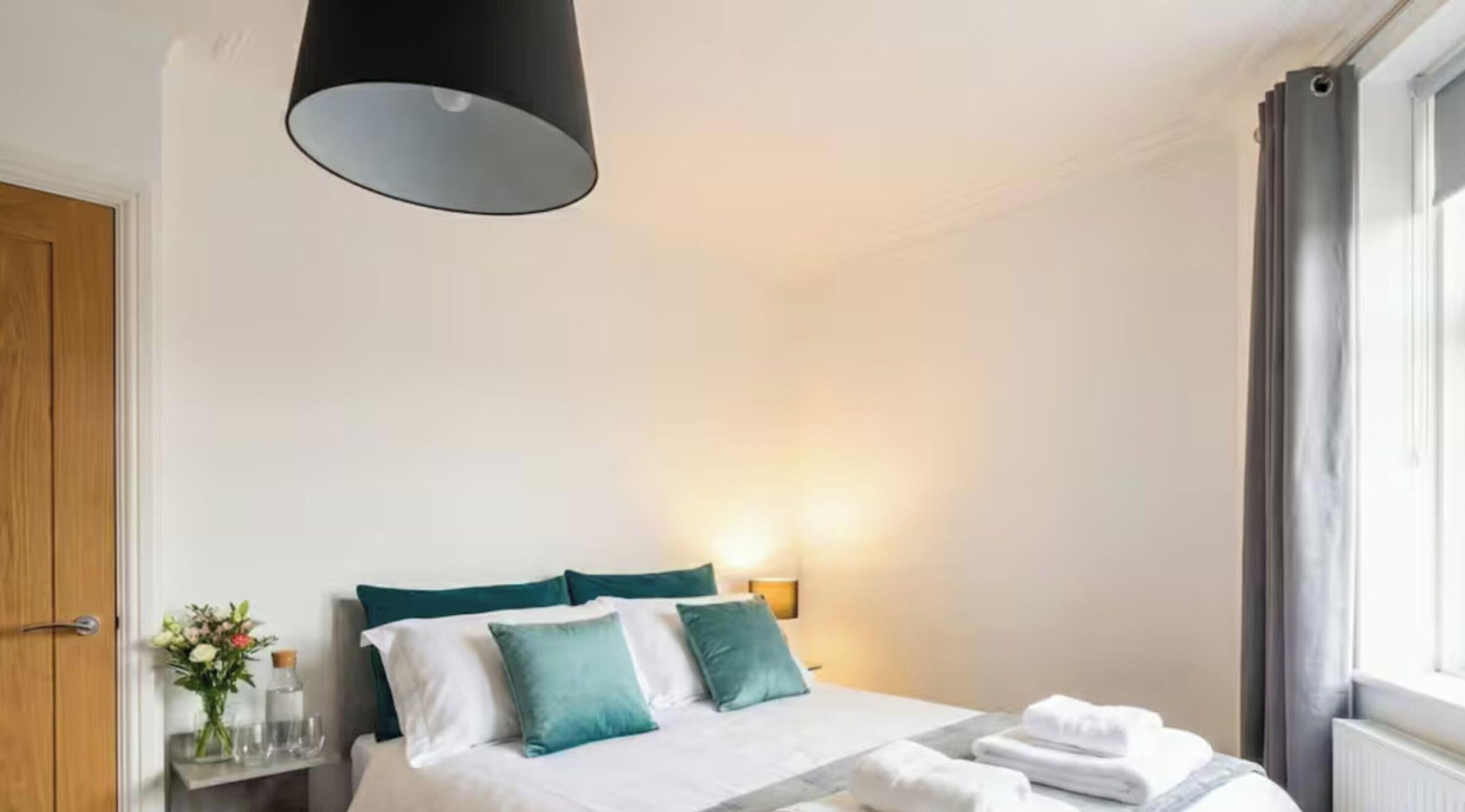 Une chambre d’hôtel avec un lit, une table de chevet ornée d’un vase de fleurs, une lampe et une fenêtre avec des rideaux.