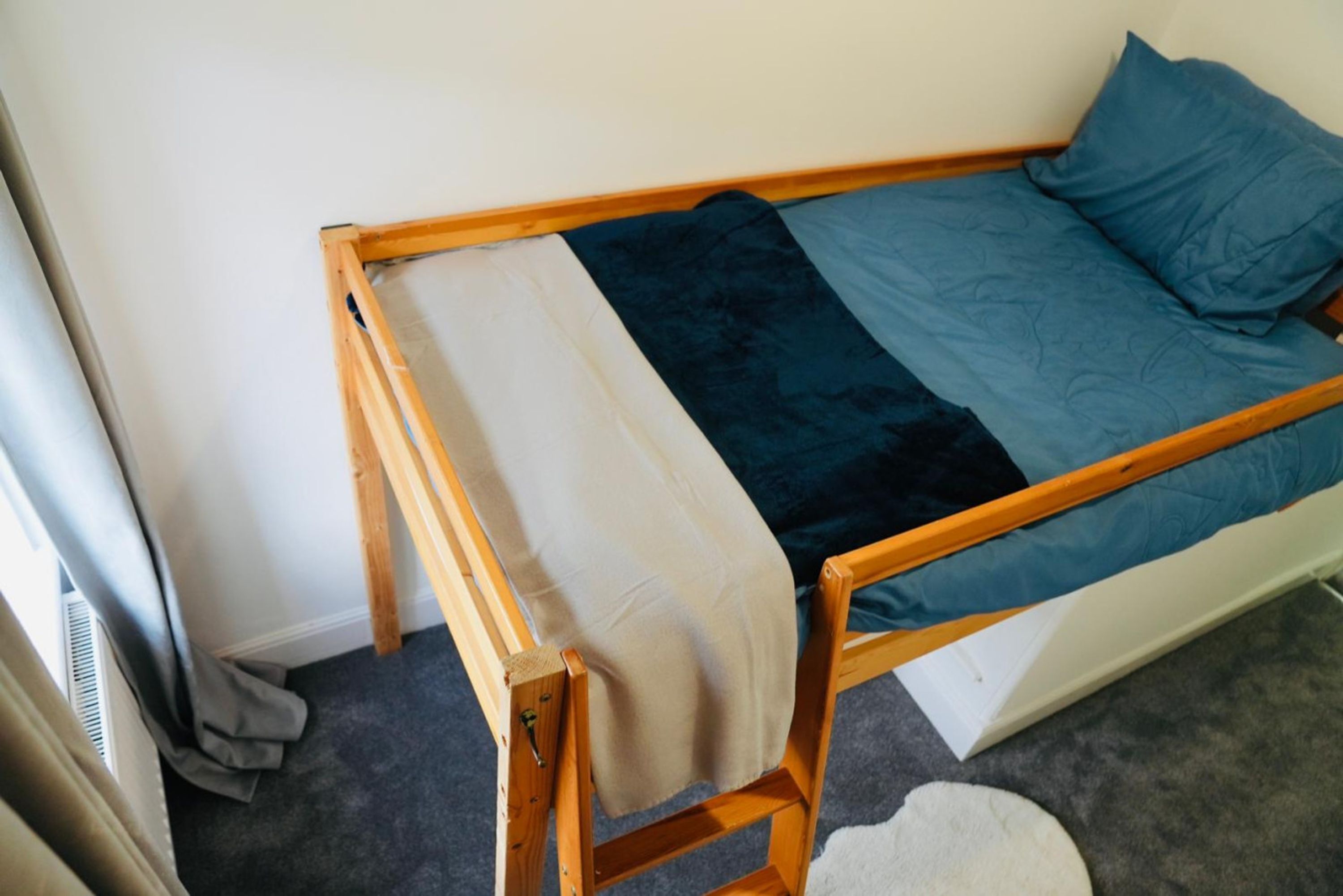 A small, single-bed room with a wooden frame, a blue mattress, and a beige blanket.
