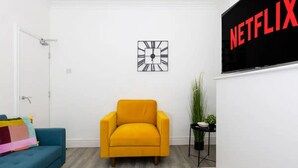 Een kamer met een blauwe bank, een gele fauteuil, een wandklok, een plant en een tv-scherm waarop 'NETFLIX' staat.