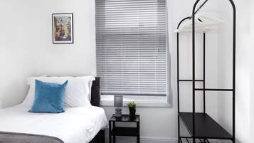 A bedroom with a bed, a nightstand, a window with blinds, and a black metal shelf unit.