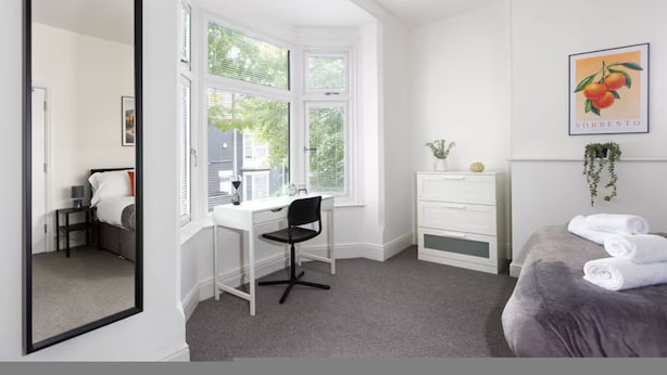 Een slaapkamer met een bed, een bureau met stoel, een dressoir, een spiegel en een raam.
