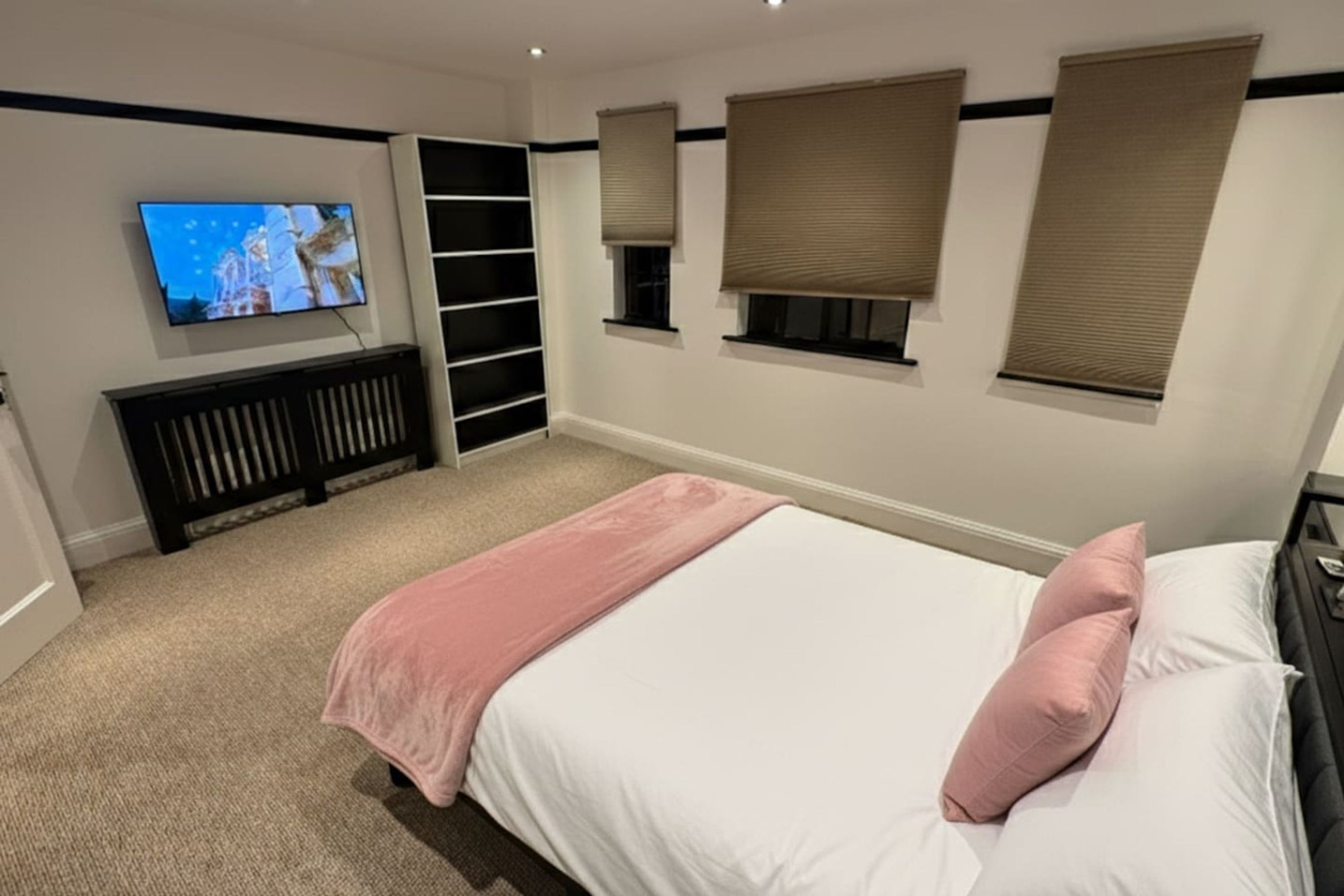 A bedroom with a bed, a TV, built-in shelving, and three windows with blinds.