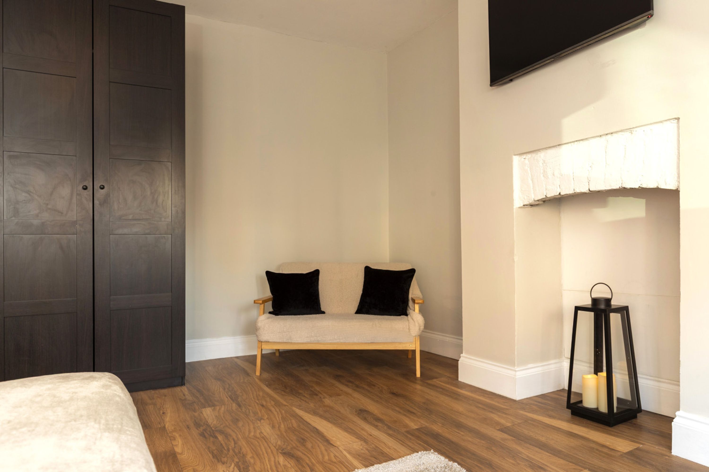 A bedroom with a bed, a dark wardrobe, a beige sofa with black cushions, a white wall-mounted lamp, and a black lantern.