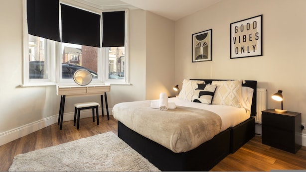 Un dormitorio con cama, mesitas de noche, un escritorio, una ventana con cortinas y dos obras de arte en las paredes.