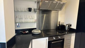 Una cocina moderna con extractor de acero inoxidable, encimera negra, horno integrado y estante de vidrio con copas y vasos.