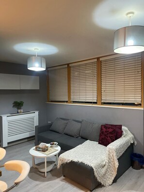 Ein kompakter Wohnbereich mit einer Couch, einem kleinen runden Tisch, einem Heizkörper und einem Fenster mit Jalousien.