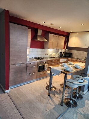A modern kitchen with wooden cabinets, a gas stove, a microwave, and a bar area with stools. - Stylish 1-bedroom Haven in Central London (London)