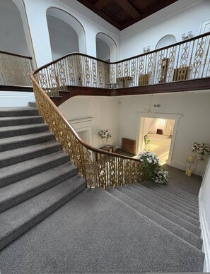 A grand staircase with ornate railings, a carpeted floor, and a view of a room with a chandelier and floral arrangement. - Room 10 - Manor Park Country House Wales (Swansea)