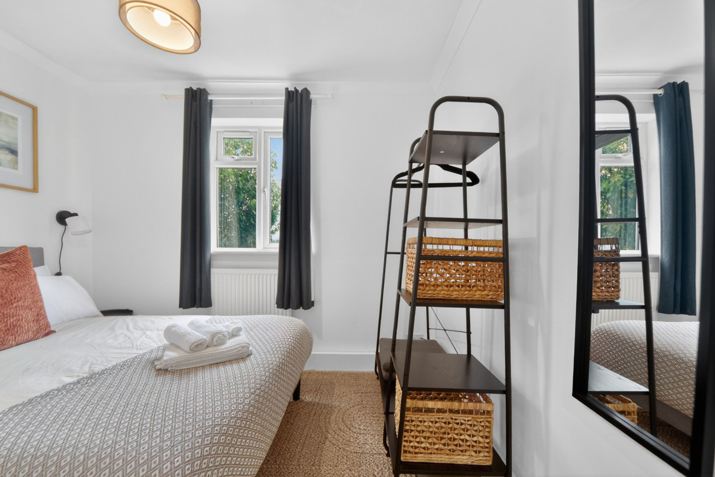 A hotel room with a bed, a window with curtains, a mirror, a ladder with baskets, and a lamp.