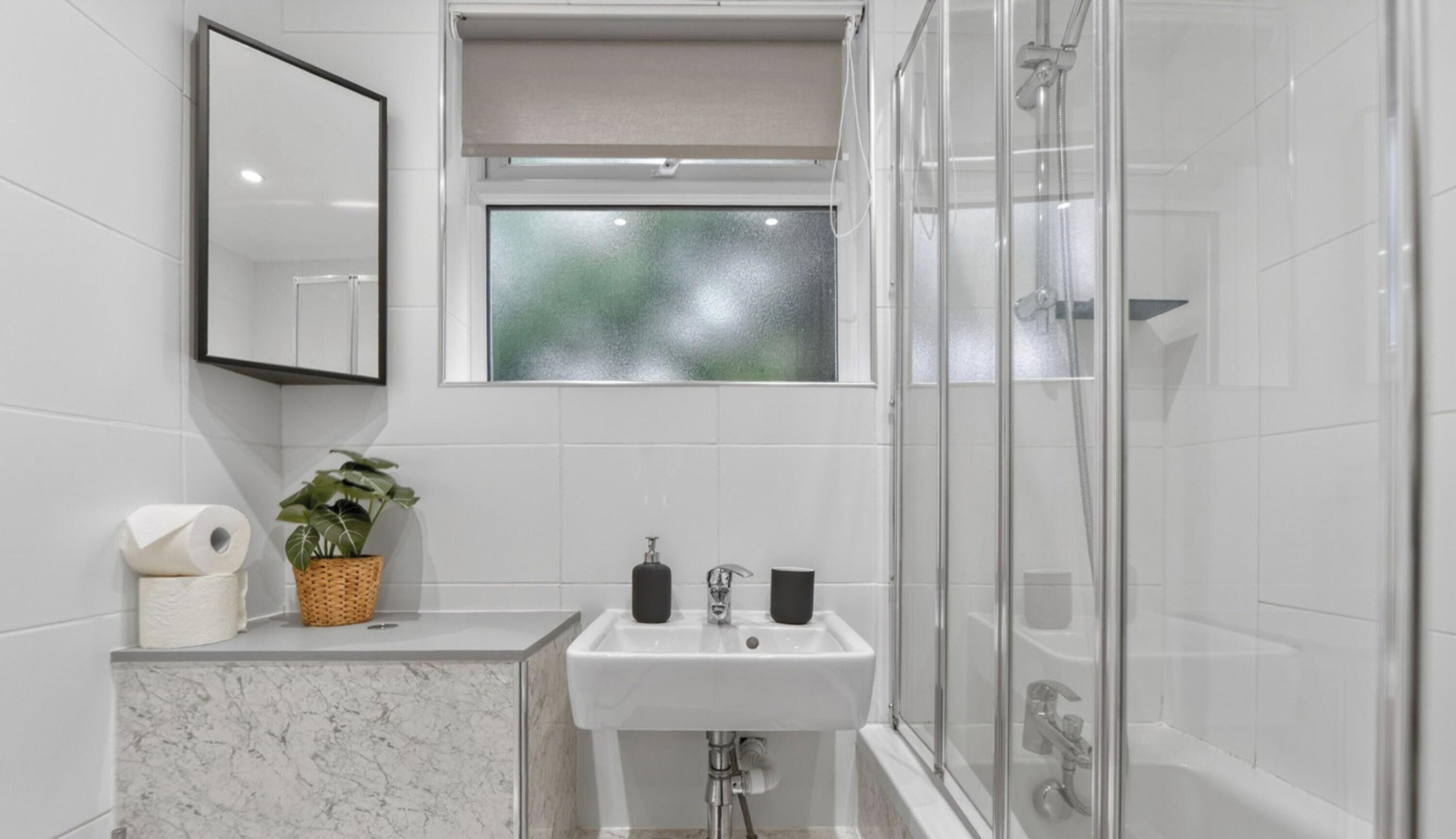 A bathroom with a marble sink, a mirror, a window, a shower, and a plant.
