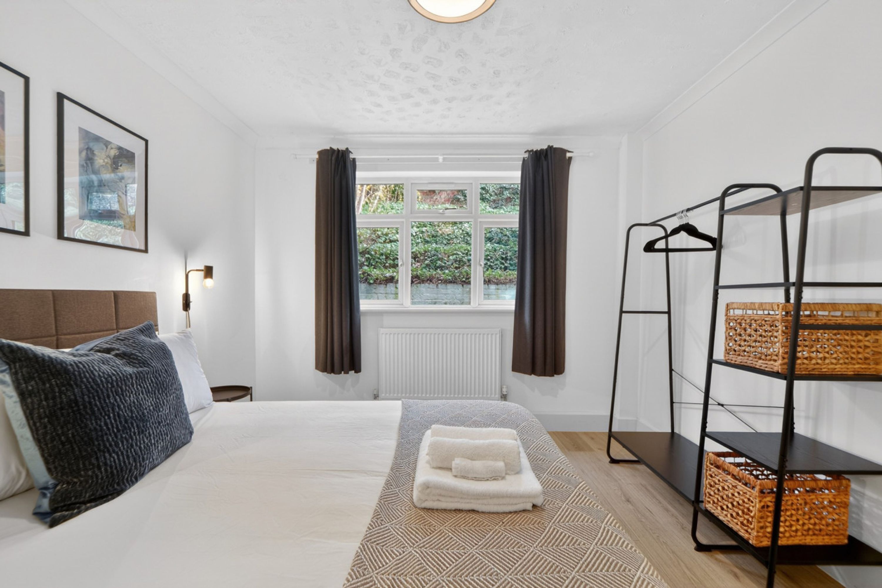 A hotel room with a bed, a window with curtains, a radiator, a wall-mounted shelf with baskets, and framed pictures on the wall.