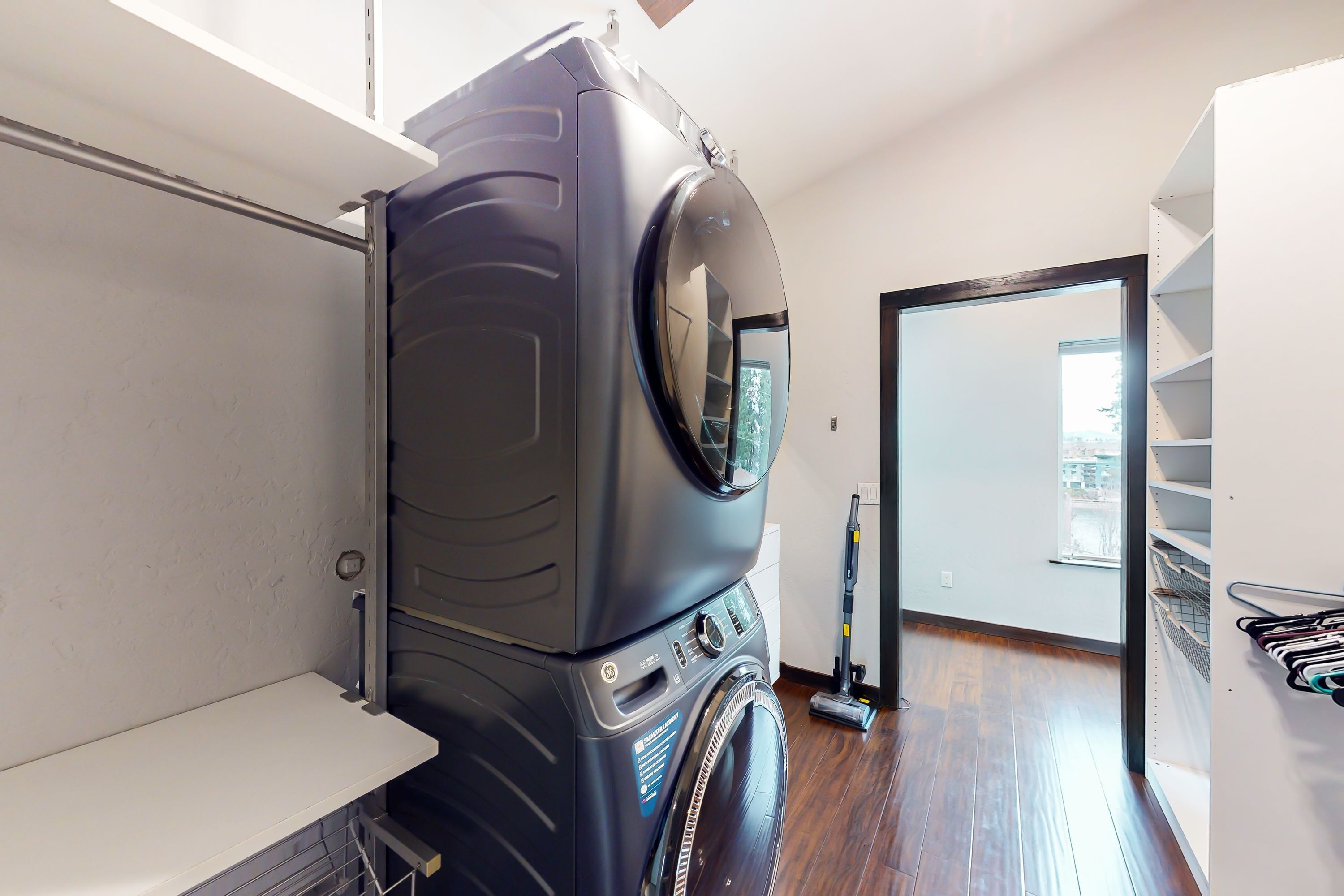 A compact laundry room with a stacked washer and dryer, a vacuum cleaner, and a shelving unit with hanging clothes.
